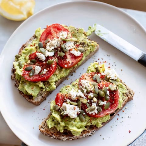 Golden toasted whole grain bread topped with mashed avocado, fresh tomato slices, and crunchy pumpkin seeds for a satisfying snack.